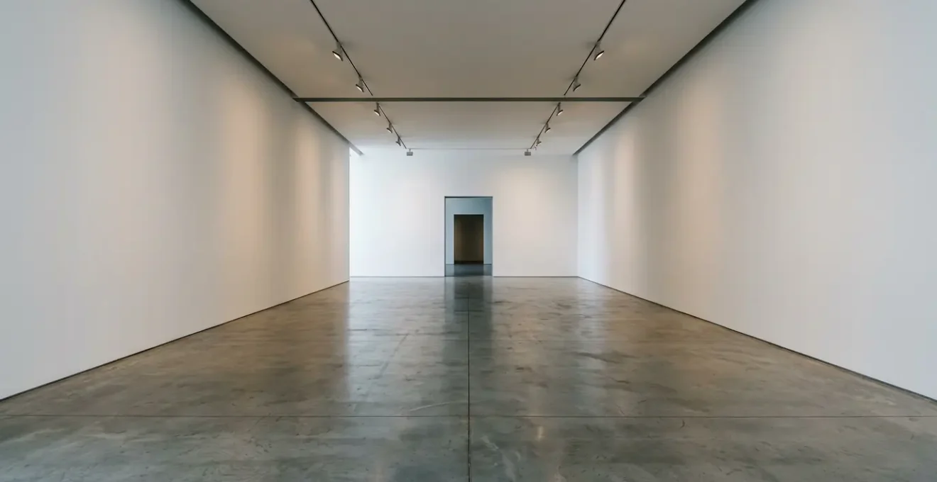 A wide-angle view of a contemporary virtual art gallery space with stark white walls and minimal lighting, emphasizing the sense of isolation and absence of human presence