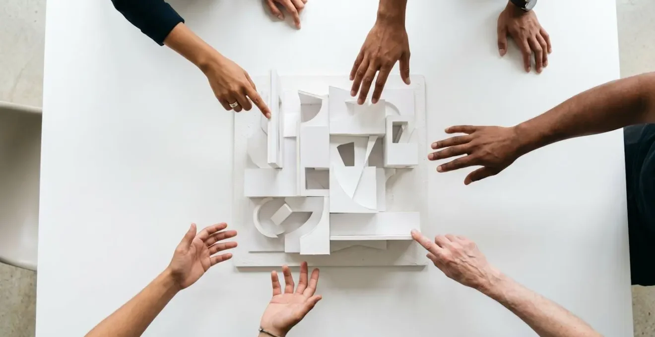 Overhead view of diverse hands pointing at and discussing abstract architectural maquette on a large table