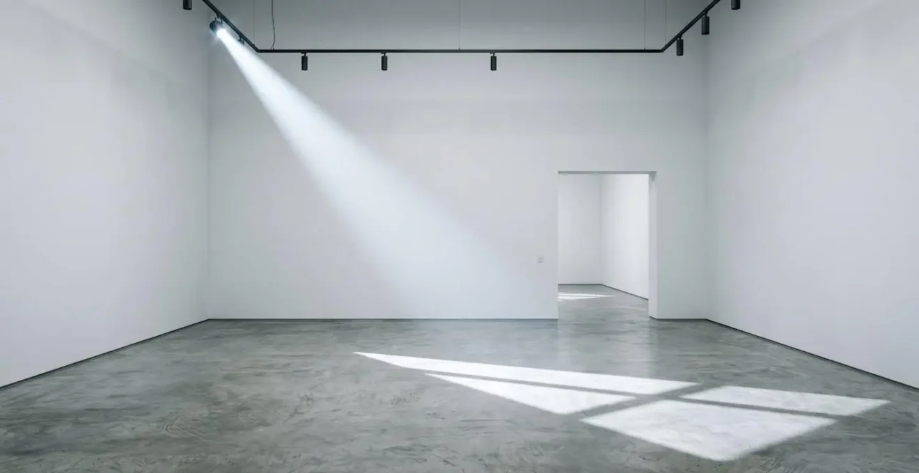 Minimalist empty white-walled gallery interior with dramatic natural lighting showing the physical overhead burden of commercial art spaces.