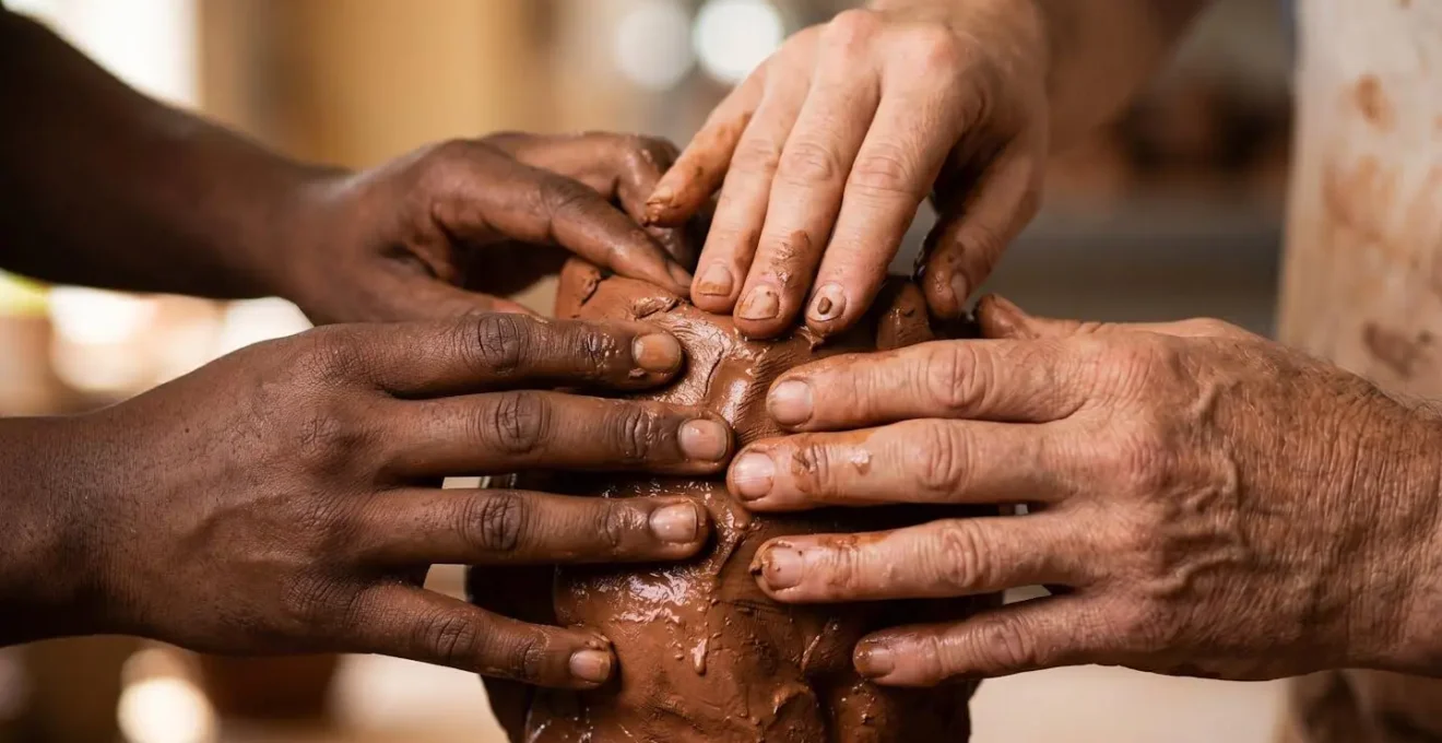 Multiple hands of different ages and skin tones collaboratively shaping wet clay on a shared pottery wheel