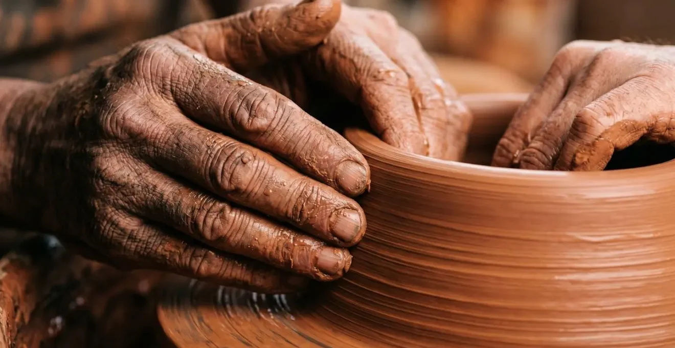 Extreme close-up of weathered craftsperson hands shaping clay on pottery wheel with visible fingerprints and texture in warm studio lighting